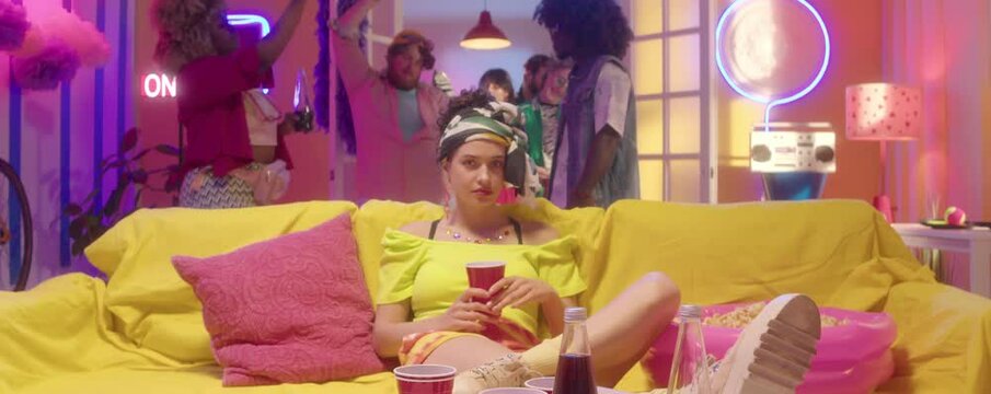 Zoom in shot of young beautiful girl in bright trendy outfit sitting alone on sofa with drink and posing on camera while friends dancing behind in party room with neon decorations