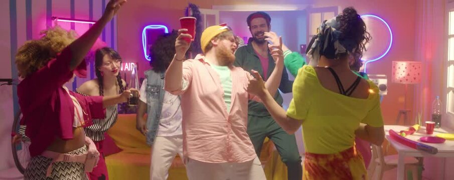 Young bearded man holding cup and dancing with company of multiethnic friends while having fun at 90s theme party in room with disco lights and neon decorations