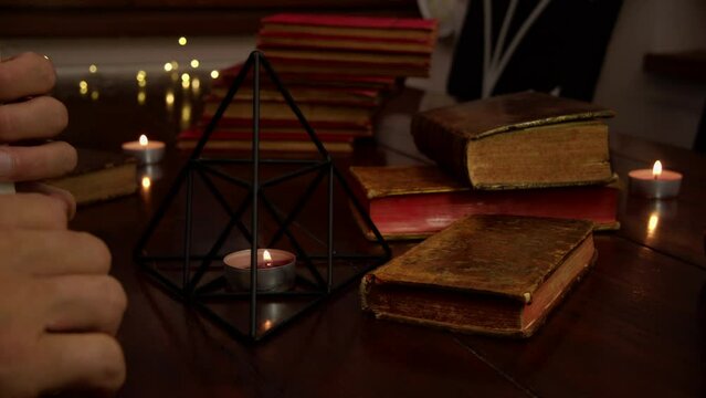 Several candles are lit on the table, an iron pyramid and antique books are visible. A man is reading and turning the pages of an antique book.