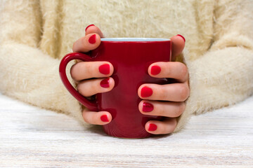 Female hands hold a red cup against the background of a white fluffy sweater with a beautiful red winter manicure. Drink, fashion, morning. New Year. Celebration. Tea coffee.