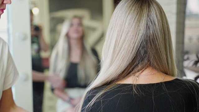 the hairdresser touches the long blond beautiful hair of a young woman in the hairdresser's chair in front of the mirror and sprays with varnish, coloring and hair care