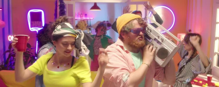 Zoom in shot of bearded man holding ghetto blaster and dancing with beautiful girlfriend while having fun at 90s theme party with friends in room with neon disco light
