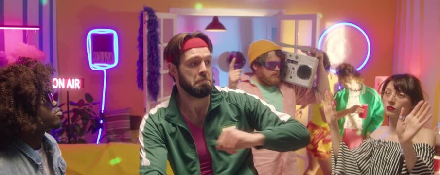 Zoom in shot of young bearded man wearing green green tracksuit and fitness headband dancing with friends at 90s disco party in room with neon light