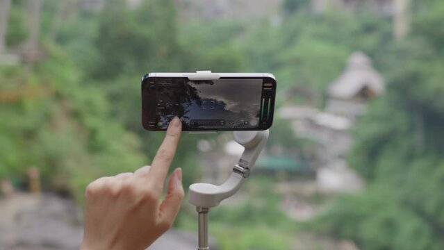 Close-up of a person changing the settings on modern phone installed on gray stabilizer against background of beautiful green panoramic landscape in defocused. video and photo shooting on the phone.