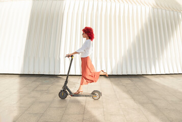 a fashionable woman with red afro hair and sunglasses rides an electric scooter with one leg...