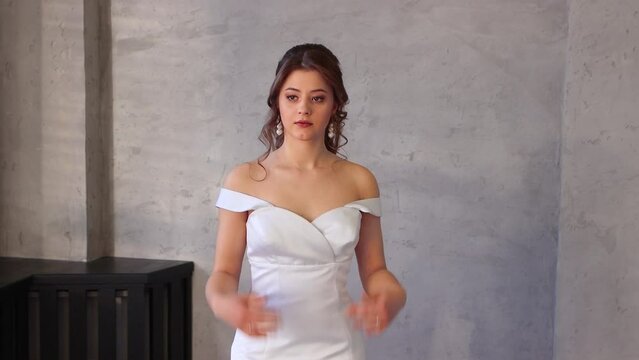 A slender beautiful girl in a white dress poses for a photographer. Sexy young brunette bride on a gray isolated background in the studio. Side copy space. UHD 4K.