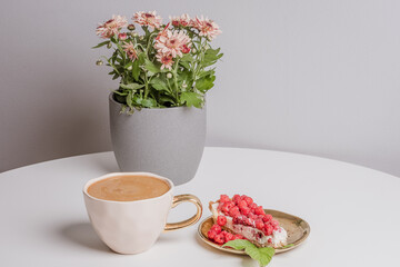 Delicious piece of cake with fresh raspberries and luxurious cup of coffee next to cake on white table. Beautiful pink chrysanthemums in grey pot in background. Light grey background. Place your text.