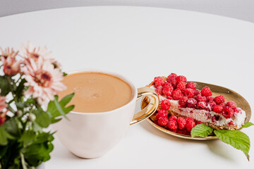 Delicious piece of cake with fresh raspberries and luxurious cup of coffee next to cake on white table. Beautiful pink chrysanthemums in grey pot. Place your text.
