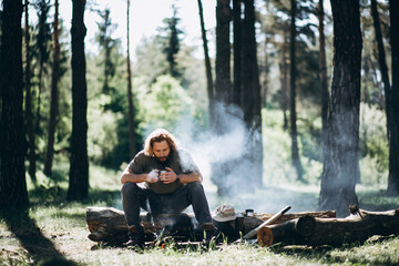 Man sits by fire in forest and drinks hot tea