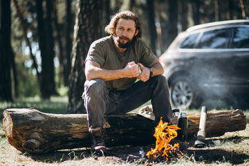 Handsome man sitting on the log by the fire in forest drinking hot tee from thermos