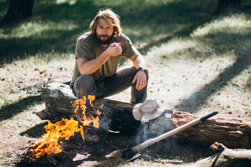 Man sits by fire in forest and drinks hot tea