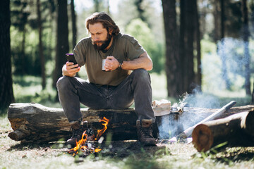 Man sits by fire in forest and drinks hot tea