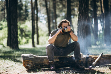 Fototapeta premium Man sits by fire in forest and drinks hot tea