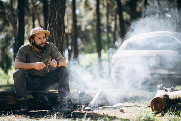 Handsome man sitting on the log by the fire in forest drinking hot tee from thermos