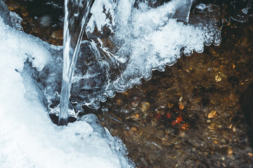 Winter stream flows through of ice and frost. Jet froze, formed icicles. Snow melting in spring in mountains.