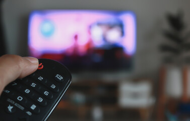 Hand holding remote control of a television. Hands pointing to tv screen set and turning it on or off select channel watching tv on his sofa at home in the living room relax.