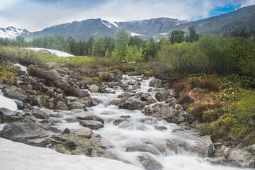 Mountain stream during spring flood, water breaks against rocks. Rivulet in mountainous area, rock ridge on horizon. Glaciers and snow on hillsides. Clouds over riverbed. - 525600709