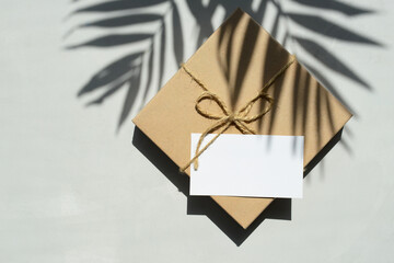 Gift box wrapped in kraft paper with blank greeting card and palm tree leaves shadows on a gray background