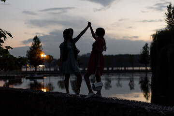 Silhouettes of two girls. Young women are holding hands against the background of the dusk sky near little lake