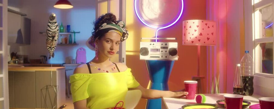 Zoom in portrait shot of beautiful brunette girl wearing bright yellow off shoulder top, trendy jewelry and headband posing on camera in room with neon decorations