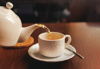 White cup with teapot. The hand holding the teapot pours tea into a pot standing on saucer in soft focus on naturally blurred background. Coffee, tea house, bokeh lights. The concept of a cozy pastime