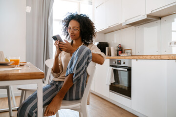 Black young woman using cellphone while having breakfast at home