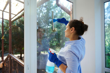 Rear view of a maid doing household chores cleaning windows, spraying glass cleanser detergent and...