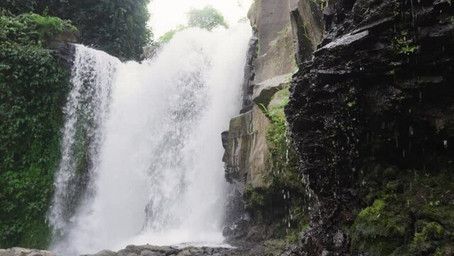A large waterfall surrounded by jungle and rocky ledges with water drops filmed in the daytime. Beautiful waterfall threshold with stone walls and tropical green foliage. Water flow in Slow motion.