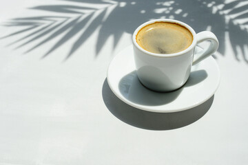 Coffee, espresso in a white coffee cup on a white table under shade of palm leaves. Black coffee and palm tree shadows on a light grey background