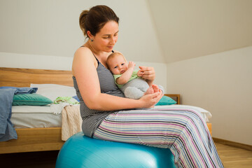 Mother holding and rocking baby sitting on fitball - obrazy, fototapety, plakaty