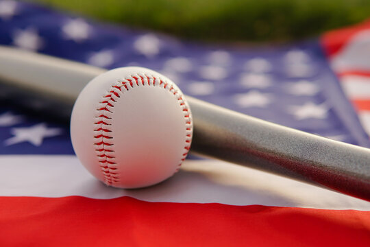 White leather ball and baseball bat laying on flag of the United States of America USA at sunset.