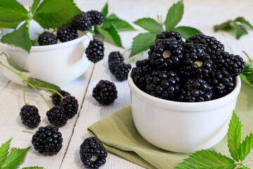 Ripe blackberries with leaves in a bowl on a whighte wooden background.