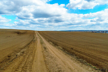 Fototapeta na wymiar A dry sandy road passes through a field under the scorching sun and clouds. Dirt road outside the city in the village. Arid climate on earth. Climate change and its consequences.