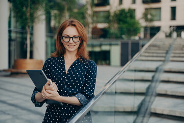 Positive redhead young woman in stylish outfit carries notepad poses outdoor