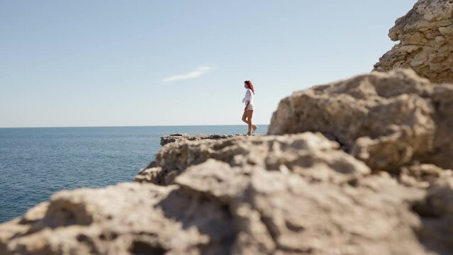 Beautiful Woman in a White Shirt and Swimsuit Walks Up to the Edge of a Cliff Against the Background of the Sea Horizon. Summer Time and Travel Concept. Go Everywhere. Slow Motion