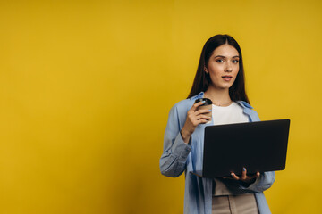 Portrait of a student girl holding a cup of coffee while working on a laptop on an isolated yellow background