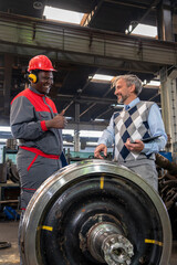 Fototapeta na wymiar Smiling Black Worker In Protective Workwear And Smiling CEO Having A Relaxed Chat. Smiling Multiracial Industrial Co-Workers Standing In A Train Factory Next To Train Wheels. 