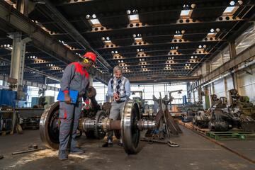 Production Manager Talking To African American Worker Next To Train Wheels At Train Factory. Area For Maintenance, Repair And Service Of Trains And Wagons. Quality Control In Manufacturing.