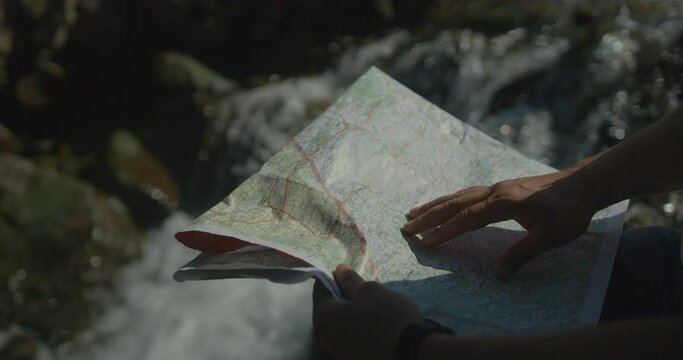 Male hands are pointing on the paper map by the finger at the background of the flowing spring in forest. Hiking and tourism concept. No face.