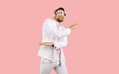 Cheerful funny young guy in a white suit, pink shirt and sunglasses with wireless pink headphones listens to music and dances on a pink studio background.