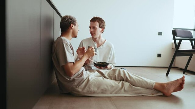 Positive gay couple eating breakfast and drinking coffee while talking on the floor in the kitchen 