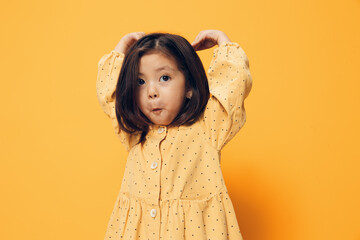 funny cute school age girl stands on an orange background with a very funny liu in a summer dress and raises her hands to her head