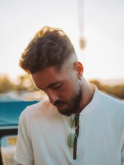 Vertical portrait of a Caucasian guy with a beard and piercing, looking down at sunset