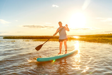 a man in shorts and a T-shirt on a SUP board with an oar floats on the water against the background of the sunset.