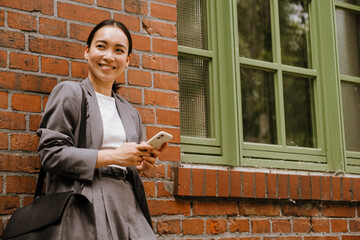 Asian businesswoman using cellphone while standing by wall outdoors