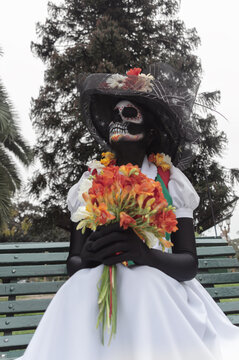 Front view of an adult woman wearing La Calavera Catrina make-up and sitting on a bench, vertical