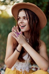 A woman in eco-friendly clothing in a hippie look sits on a colored plaid smiling and looking at an autumn sunset in nature in the park