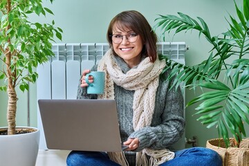 Woman in warm clothes sitting near heating radiator, using laptop