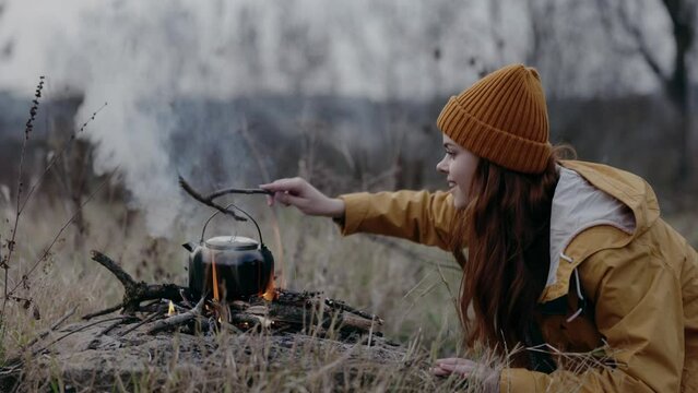 A woman in a yellow hat in a camping trip blows up a fire in the countryside to boil water in a kettle to keep warm in the fall on a trip