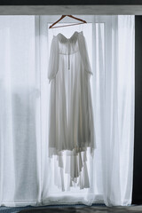 A beautiful long white bridesmaid dress hangs on a wooden hanger in front of the window. wedding photography.